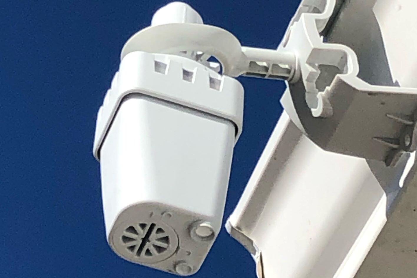 A rain sensor, a white container with a grid at the bottom is attached to the edge of a building outside.