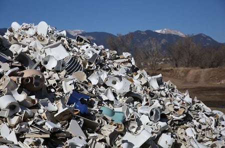 A large pile of broken toilets to be recycled. The Rocky Mountains are visible on the horizon in the background. The sky is blue and cloudless.