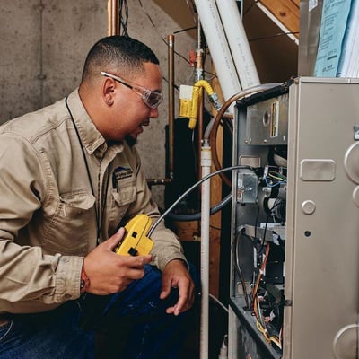 A utility worker wears safety glasses and a beige shirt kneels in front of an open electrical panel on a furnace. He's holding a yellow tool.