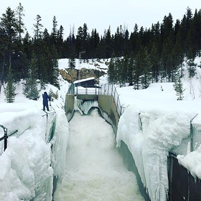 Water flows rapidly from a large mountain tunnel. Snow and ice are visible on both sides of the spillway. A person in a blue coat watches nearby.