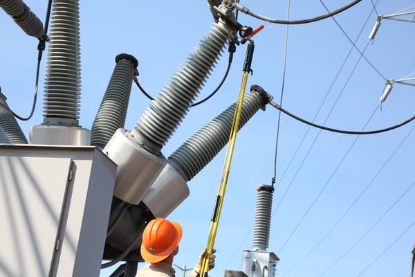 A utility worker wearing safety gear uses a tool on a yellow pole to repair a large electrical transformer at an outdoor power station.