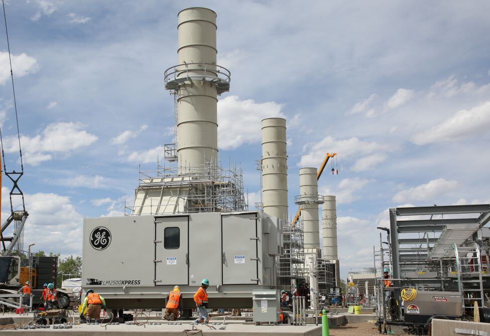 Installation of our new Natural Gas Generators. Workers in safety gear stand near a grey building near a smokestack. Four total stacks are visible.