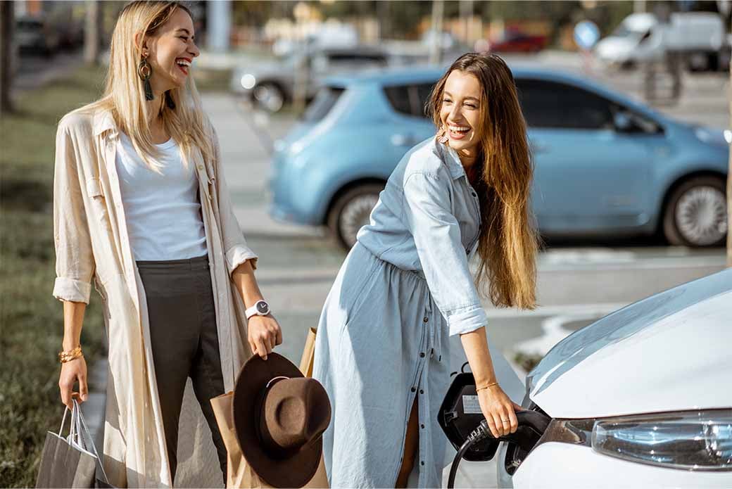 Two women stand smiling next to a white electric car parked outside. One plugs a charging cable into the car, the other is holding shopping bags. 