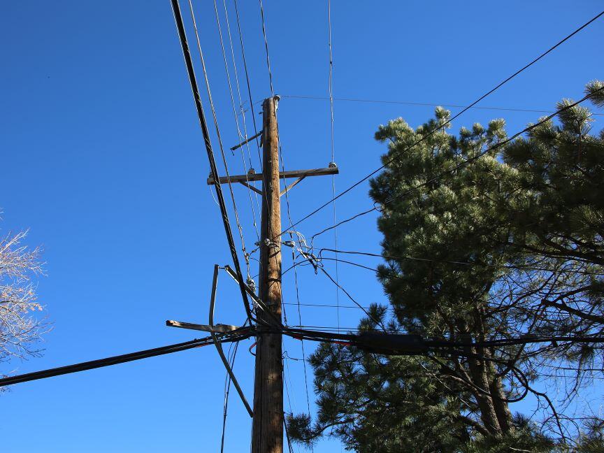 Photo displays a wooden utility pole with wires and cables that were damaged in the December 2021 windstorm, set against a clear blue sky. 