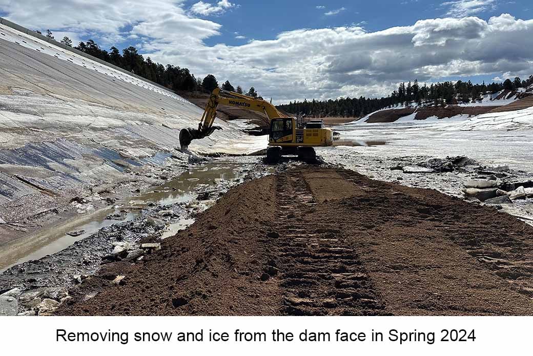 Excavator claw scrapes ice from a steel dam face. Muddy tracks are visible. Text reads "Removing snow & ice from the dam face in Spring 2024"