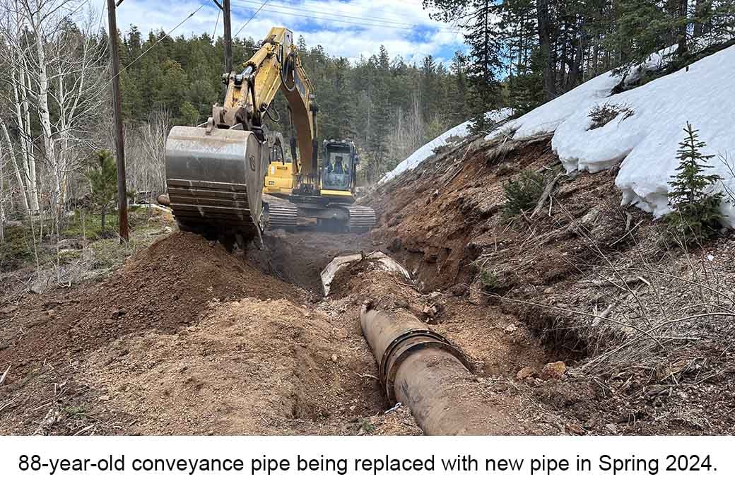 Excavator digs near a large pipe in a forest. Text reads: "88-year-old conveyance pipe being replaced with new pipe in Spring 2024."