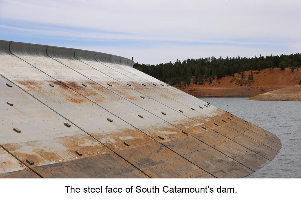 Grey curved steel dam face next to low water. A tree-lined shore is in the distance. Text overlay reads: "The steel face of South Catamount's dam"