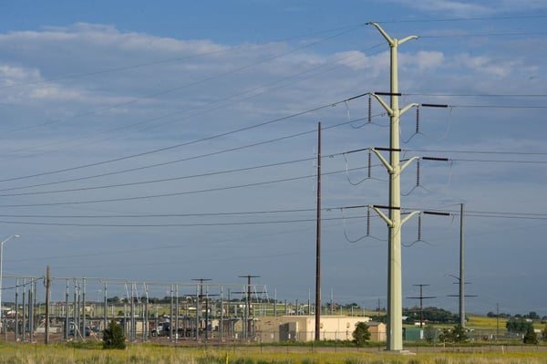 A large metal power pole with crossbeams and suspended power lines. An industrial facility with small structures & a fence is visible in the distance