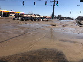 Muddy water covers the intersection of Maizeland Road and Academy Boulevard after a water main break. Several cars are stopped near a gas station.