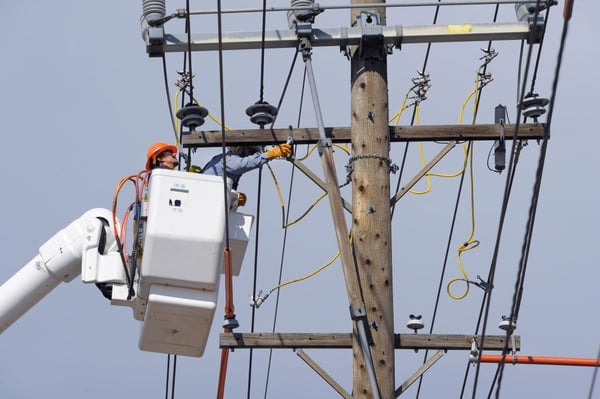 A worker in safety gear in an elevated bucket performs maintenance on electric lines near a wooden utility pole.