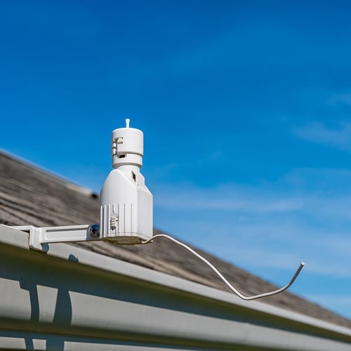 A rain sensor, a white container with a wire sticking out at the bottom is attached to the edge of a building outside.