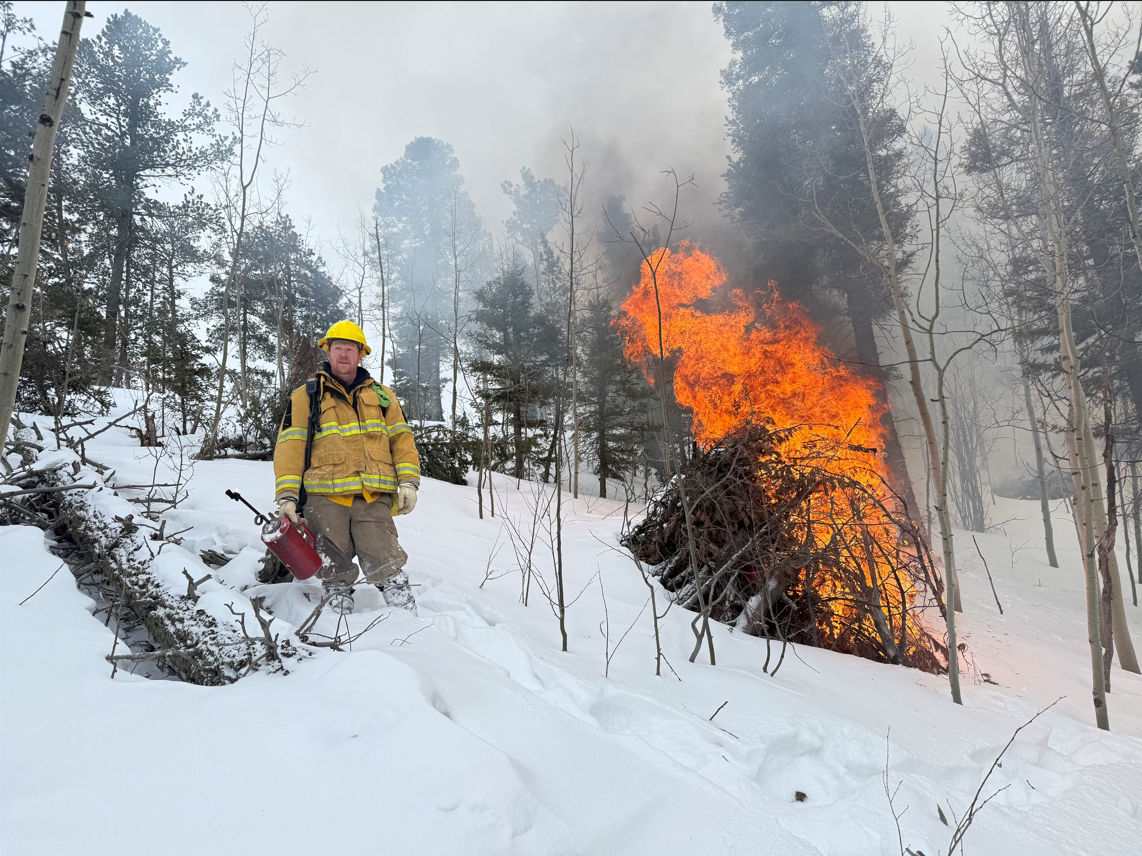 A firefighter standing in snow next to a pile of brush on fire.