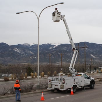 Two workers wearing safety gear repair a streetlight on a road in Colorado Springs. One worker is in a bucket lifted above a truck next to a streetlight. The other is on the road next to construction cones.