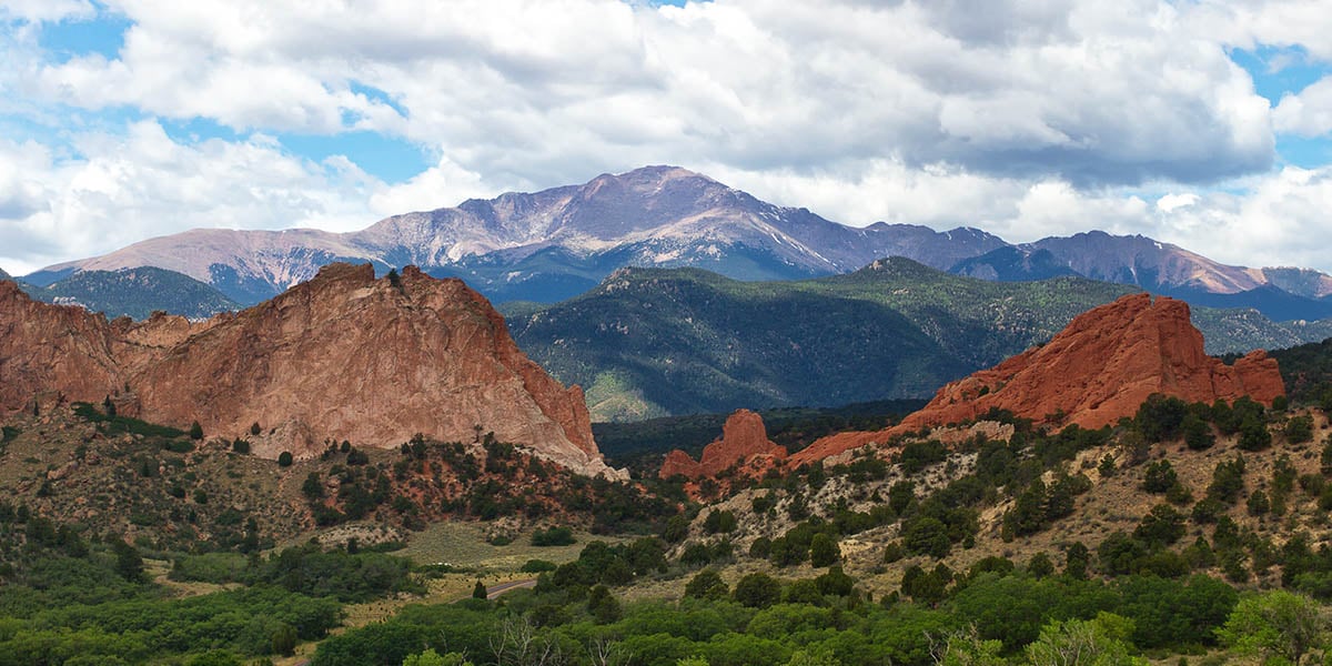 Pikes Peak towers behind the red rock formations of Garden of the Gods on a partly cloudy day.