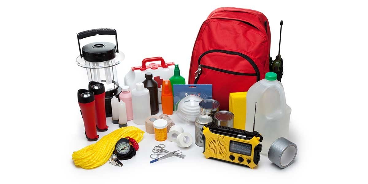 Emergency supplies including water, a radio, various medical supplies, flashlights, and canned food sit on the ground with a red backpack.