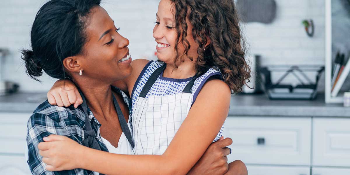 A mother and daughter smiling wearing aprons embracing in a kitchen. 